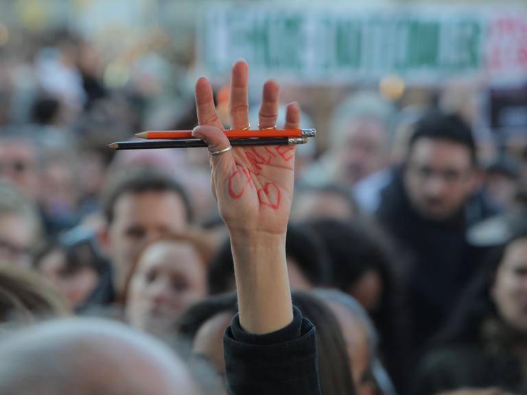 Eine Person in einer Menschenmenge hält ihre Hand nach oben, in der sie zwei Stifte hält. Auf ihrer Hand steht "Charlie" geschrieben und daneben ist ein Herz gezeichnet. 