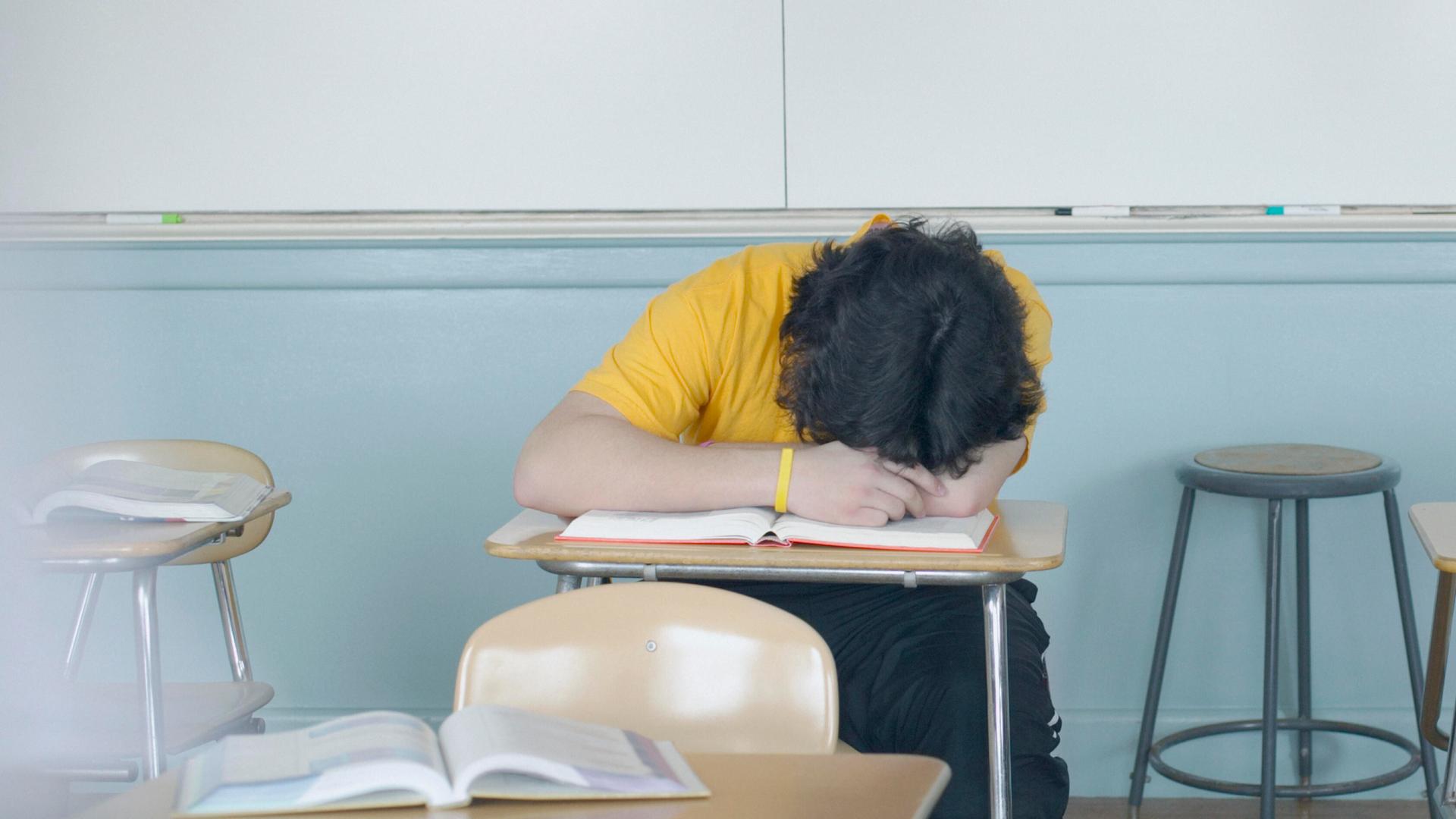 In einem Klassenzimmer liegt ein Teenager im gelben T-Shirt mit dem Kopf auf dem Tisch. In einem Klassenzimmer liegt ein Teenager im gelben T-Shirt mit dem Kopf auf dem Tisch.