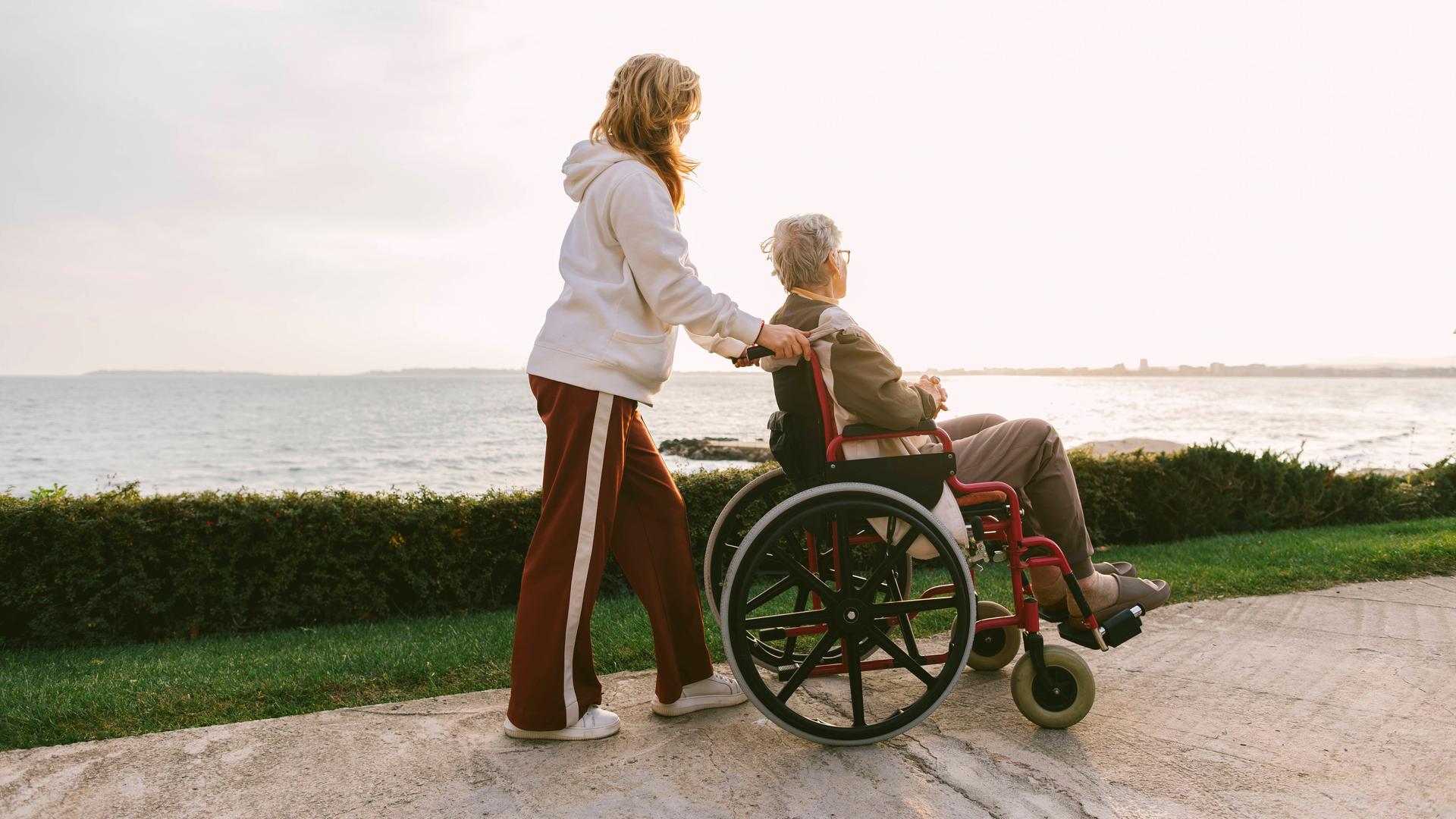 Eine Frau hilft einer anderen Frau im Roll-Stuhl. Sie sind auf einem Küsten-Weg.