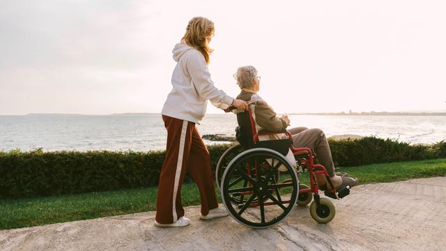 Eine Frau hilft einer anderen Frau im Roll-Stuhl. Sie sind auf einem Küsten-Weg.