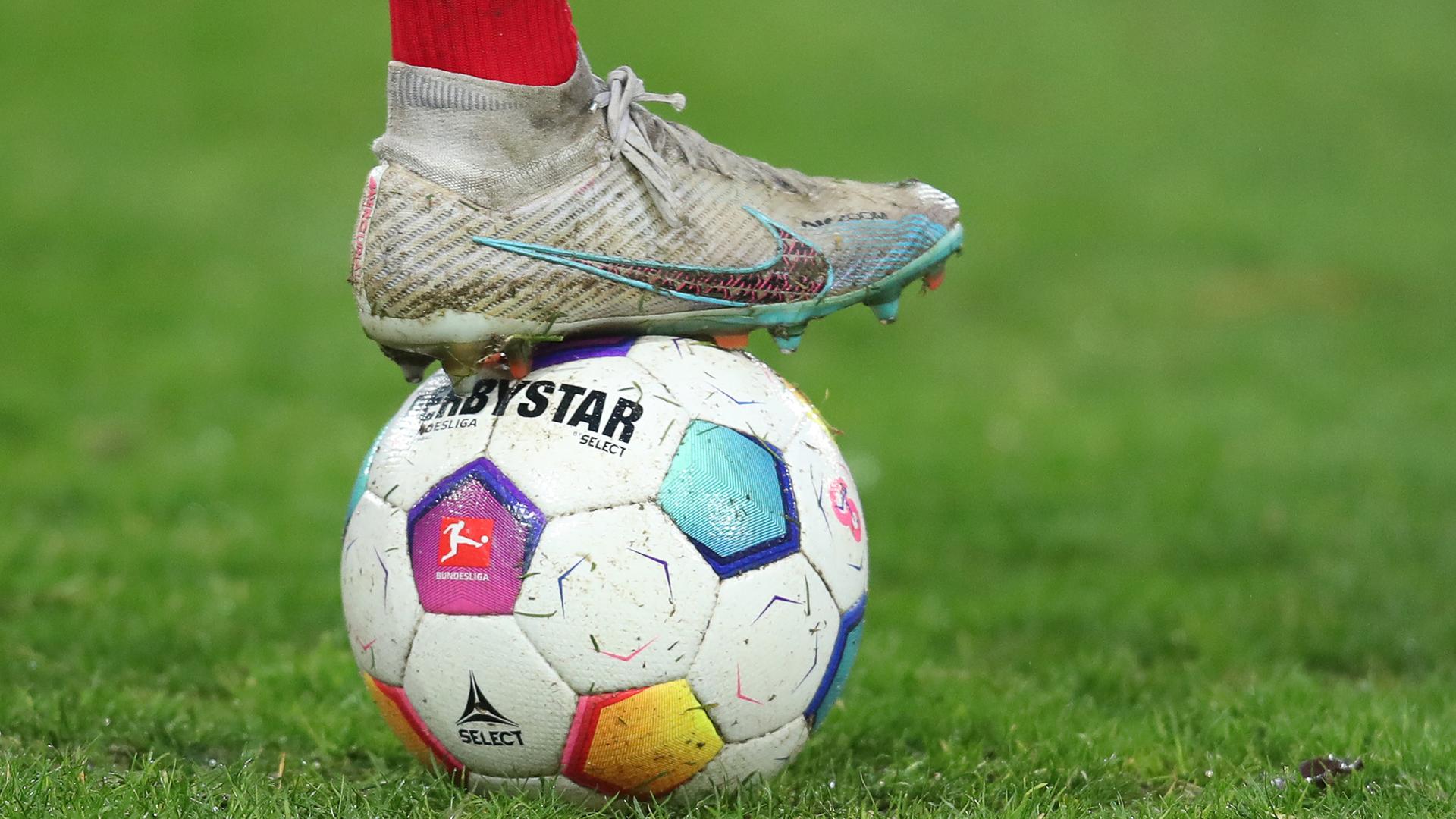 Ein weiß-bunter Profifußball wird von einem Spielerfuß in Sportschuhen festgehalten.