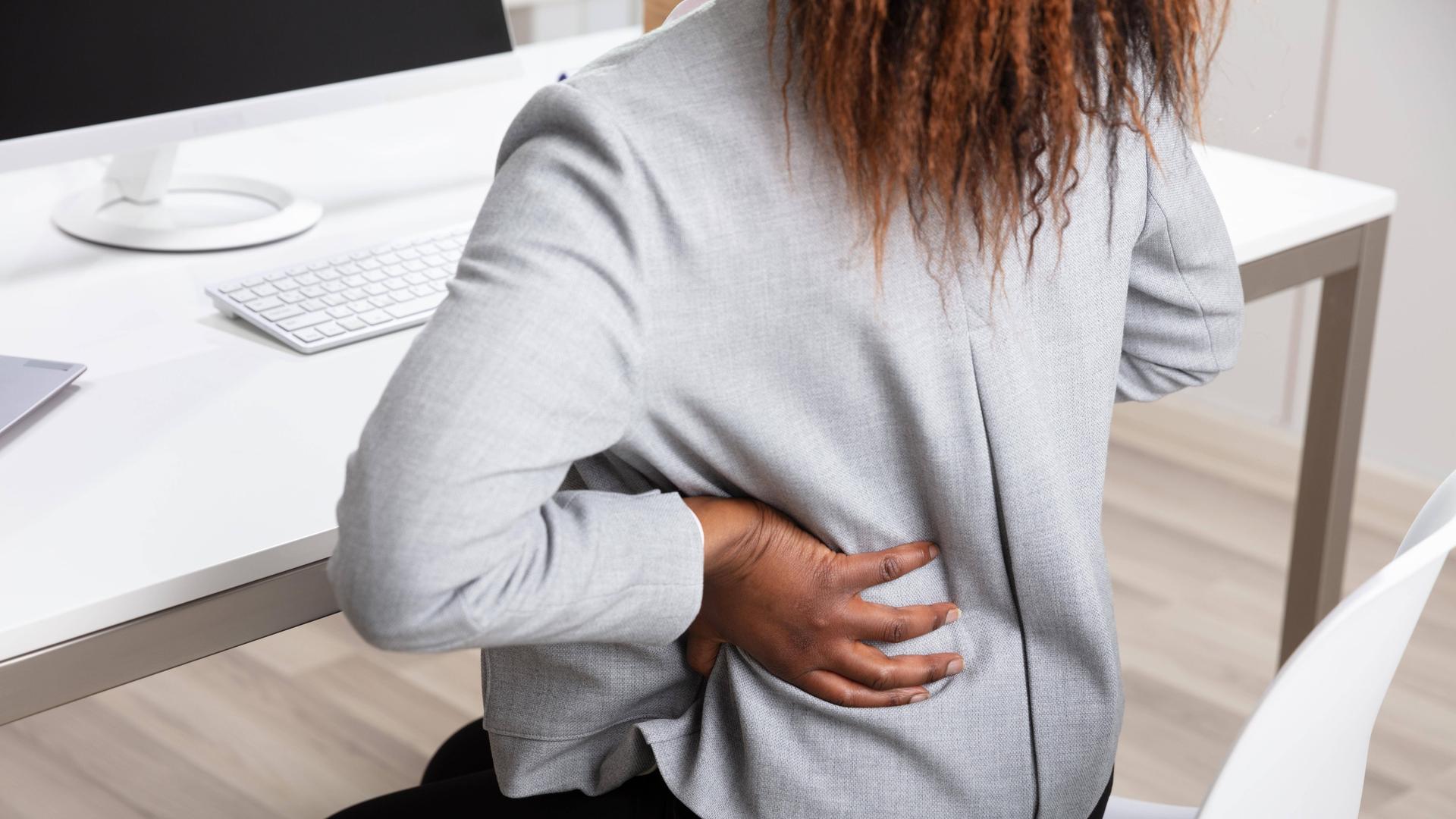Eine Frau sitzt an einem Schreibtisch und hält ihre Hand an den Rücken, als habe sie Schmerzen. 