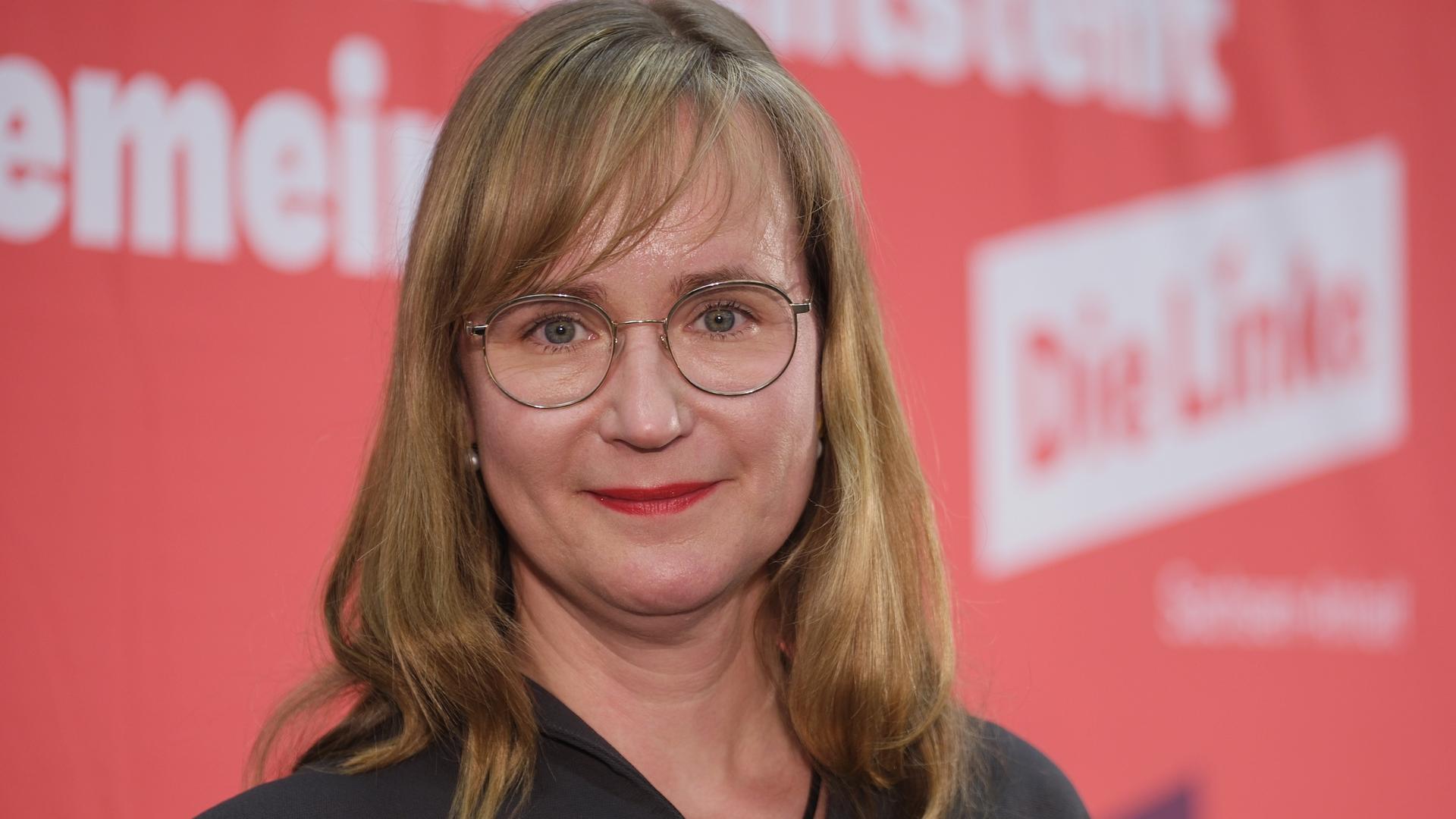 Eine blonde Frau mit Brille lächelt in die Kamera. Im Hintergrund ist der Schriftzug "Die Linke" zu sehen.