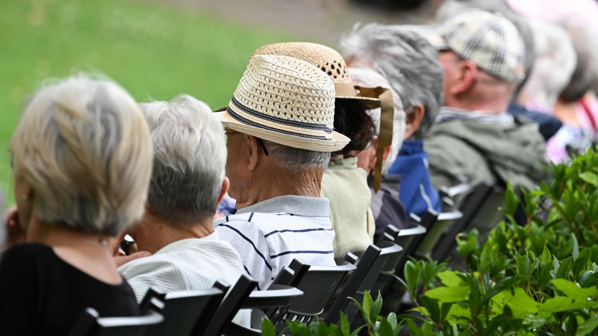 Mehrere ältere Menschen sitzen in einem Kurzpark auf Stühlen in einer Reihe. Man sieht ihre Köpfe von seitlich-hinten, ihre Gesichter sind nicht zu erkennen. 