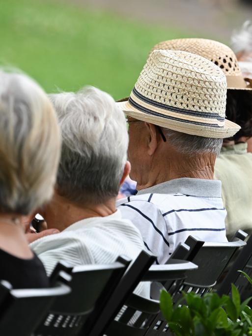 Mehrere ältere Menschen sitzen in einem Kurzpark auf Stühlen in einer Reihe. Man sieht ihre Köpfe von seitlich-hinten, ihre Gesichter sind nicht zu erkennen. 