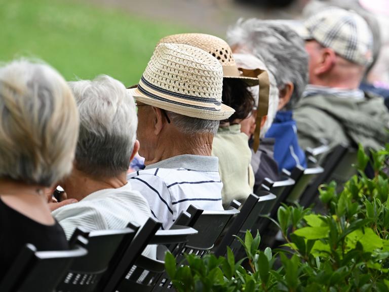 Mehrere ältere Menschen sitzen in einem Kurzpark auf Stühlen in einer Reihe. Man sieht ihre Köpfe von seitlich-hinten, ihre Gesichter sind nicht zu erkennen. 