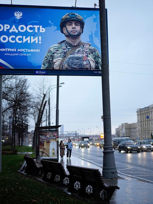 Zwei Frauen gehen unter einem großen Werbeplakat für das russische Militär lang.