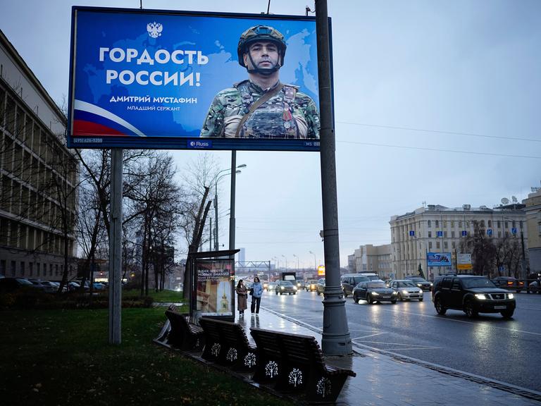 Zwei Frauen gehen unter einem großen Werbeplakat für das russische Militär lang.