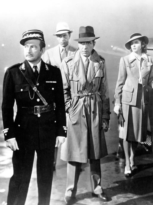 Casablanca. Mehrere Personen stehen bei Regenwetter auf einem Platz. Zwei Personen in Uniform, drei weitere Personen in Zivilkleidung. 