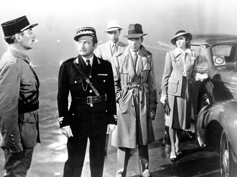 Casablanca. Mehrere Personen stehen bei Regenwetter auf einem Platz. Zwei Personen in Uniform, drei weitere Personen in Zivilkleidung. 