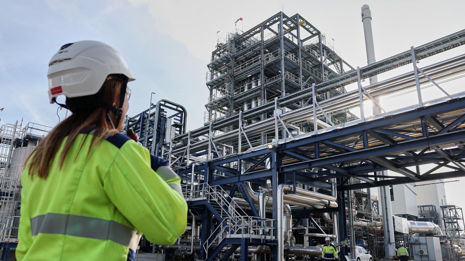 Eine Mitarbeiterin der UPM-Bioraffinerie Leuna spricht in ihr Funkgerät. Im Hintergrund ist ein hoher Turm aus Stahlrohren zu sehen. Eine Mitarbeiterin der UPM-Bioraffinerie Leuna spricht in ihr Funkgerät. Im Hintergrund ist ein hoher Turm aus Stahlrohren zu sehen.