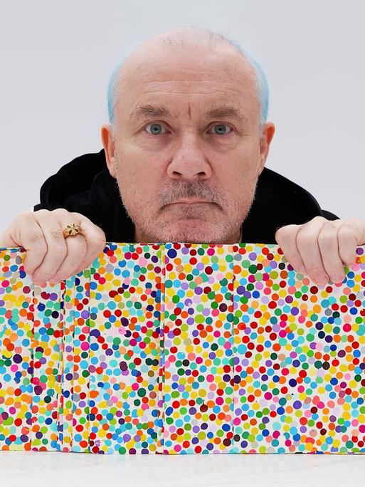 Damien Hirst mit Kunstwerken seiner „Currency“ Reihe in der Hand, Bildern mit vielen Punkten.