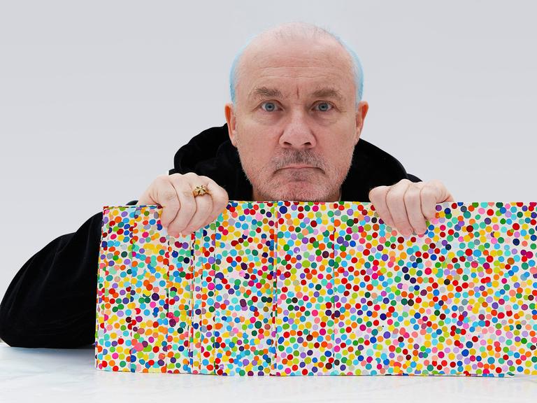 Damien Hirst mit Kunstwerken seiner „Currency“ Reihe in der Hand, Bildern mit vielen Punkten.