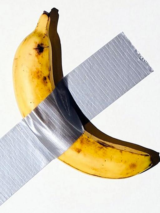 Eine reife Banane mit kleinen braunen Flecken klebt unter einem grauen Klebestreifen an einer Wand.