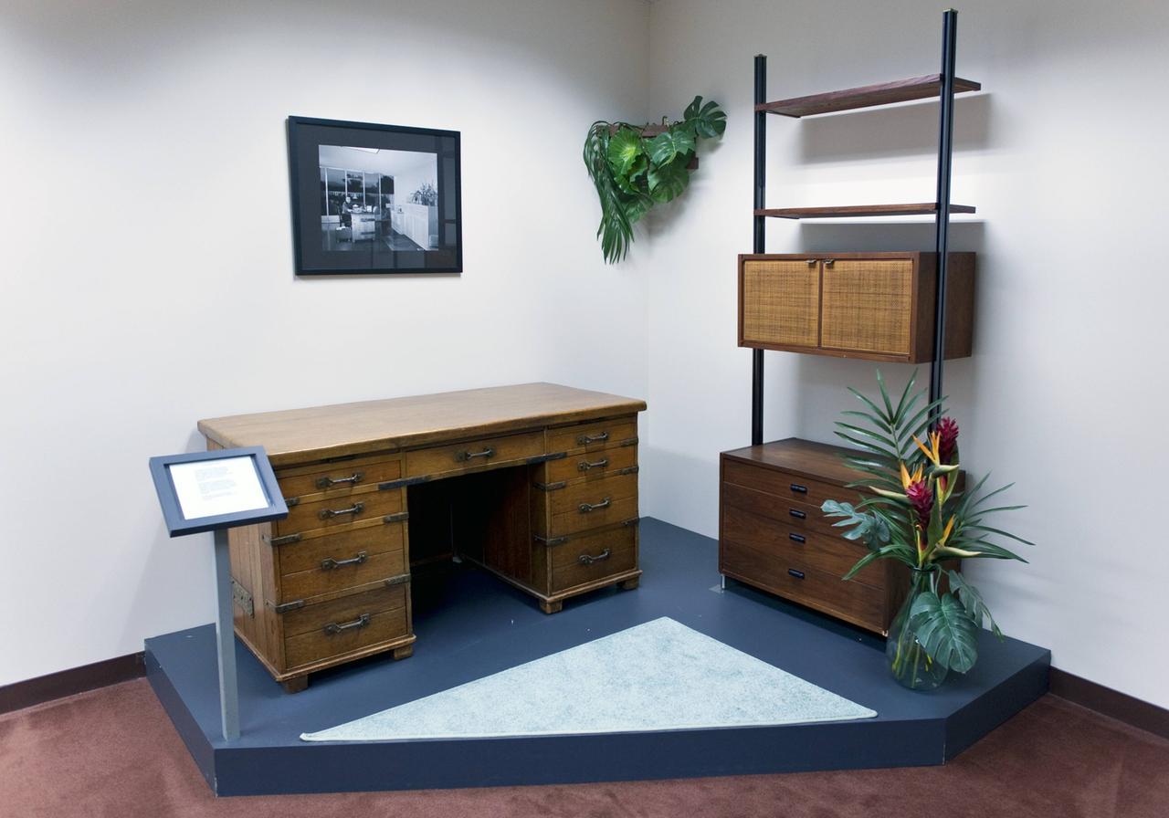 Ein hölzerner Schreibtisch mit Schubladen ist im Museum auf einem Podest ausgestellt. Regale und Zimmerpflanze komplettieren den Ausschnitt einer Büroeinrichtung.