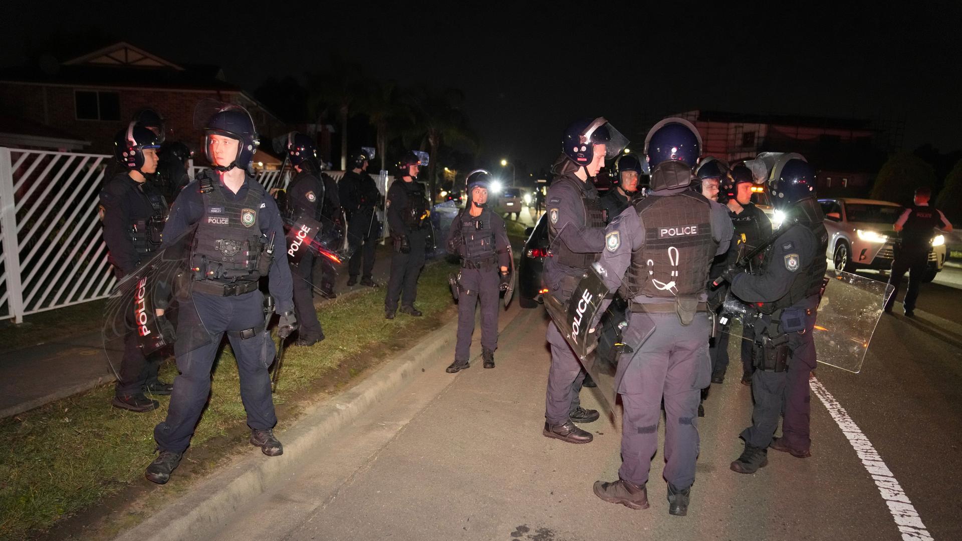 Es ist dunkel: Mehre bewaffnete Polizisten in dunkleblauen Uniformen mit Helmen und Schildern und stehen an einer beleuchteten Straße.