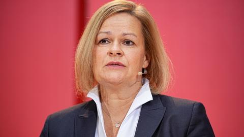 Nancy Faeser mit dunklem Jackett und weißem Hemd vor einer roten Wand.