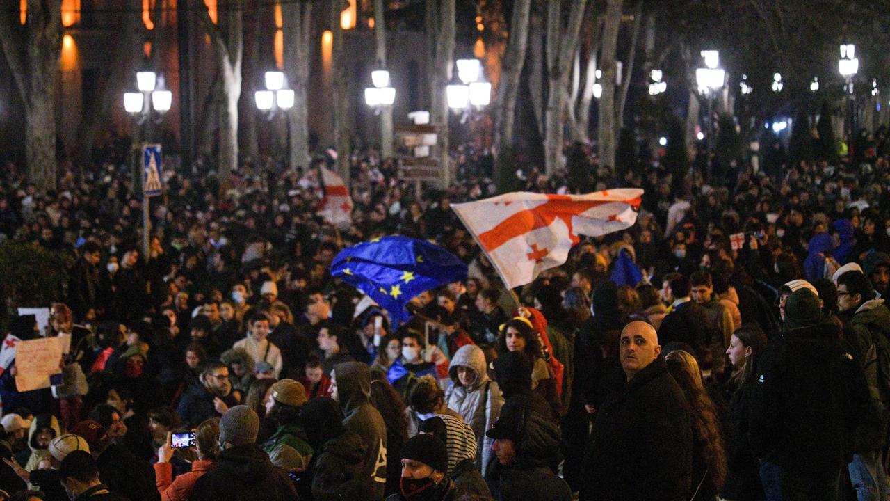 Auf einer Straße in der georgischen Hauptstadt Tbilisi stehen im März 2023 im Dunkeln zahlreiche Protestierende, eine Georgien- und eine EU-Flagge sind zu sehen. 