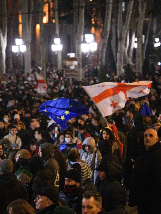 Auf einer Straße in der georgischen Hauptstadt Tbilisi stehen im März 2023 im Dunkeln zahlreiche Protestierende, eine Georgien- und eine EU-Flagge sind zu sehen. 