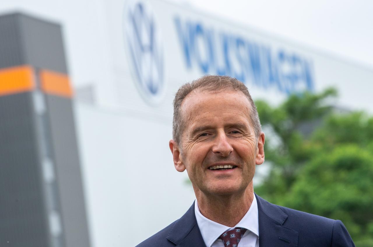 Porträt von Herbert Diess. Im Hintergrund steht an einer Halle der Schriftzug von Volkswagen.