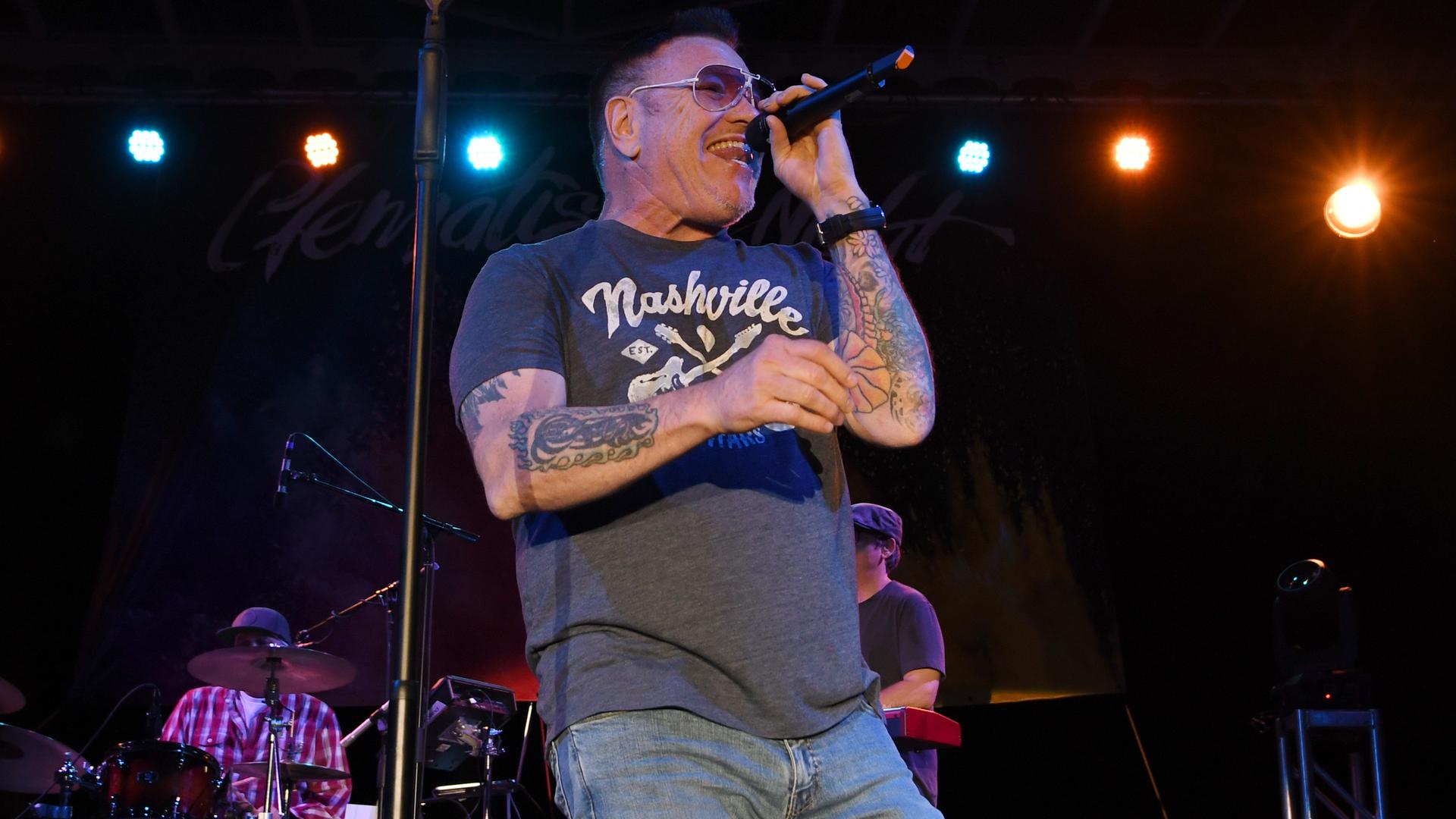 Harwell im grauen Shirt mit "Nashville"-Aufdruck und Sonnenbrille singt auf der Bühne. Hinter ihm ein Keyboarder und ein Schlagzeuger. Harwell im grauen Shirt mit "Nashville"-Aufdruck und Sonnenbrille singt auf der Bühne. Hinter ihm ein Keyboarder und ein Schlagzeuger.