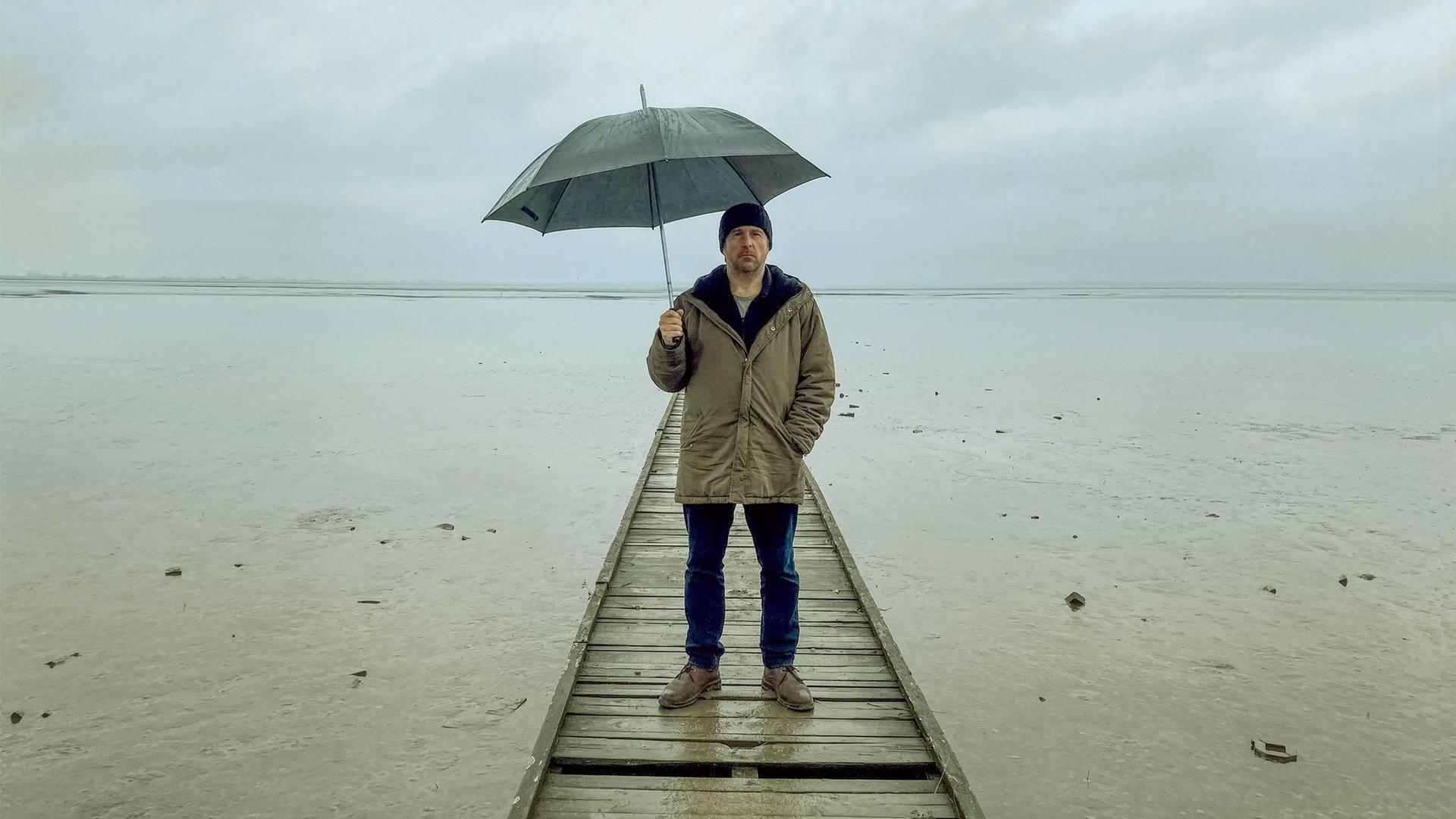 Bjarne Mädel als Kriminalkommissar Sörensen auf einem Steg im Wasser, einen Regenschirm über sich haltend.