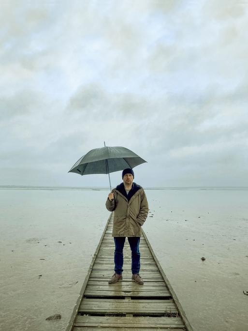 Bjarne Mädel als Kriminalkommissar Sörensen auf einem Steg im Wasser, einen Regenschirm über sich haltend.