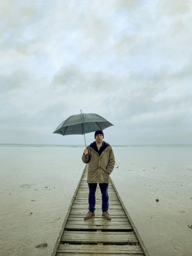 Bjarne Mädel als Kriminalkommissar Sörensen auf einem Steg im Wasser, einen Regenschirm über sich haltend.