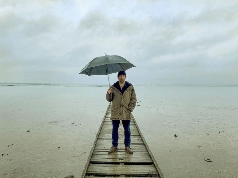 Bjarne Mädel als Kriminalkommissar Sörensen auf einem Steg im Wasser, einen Regenschirm über sich haltend.