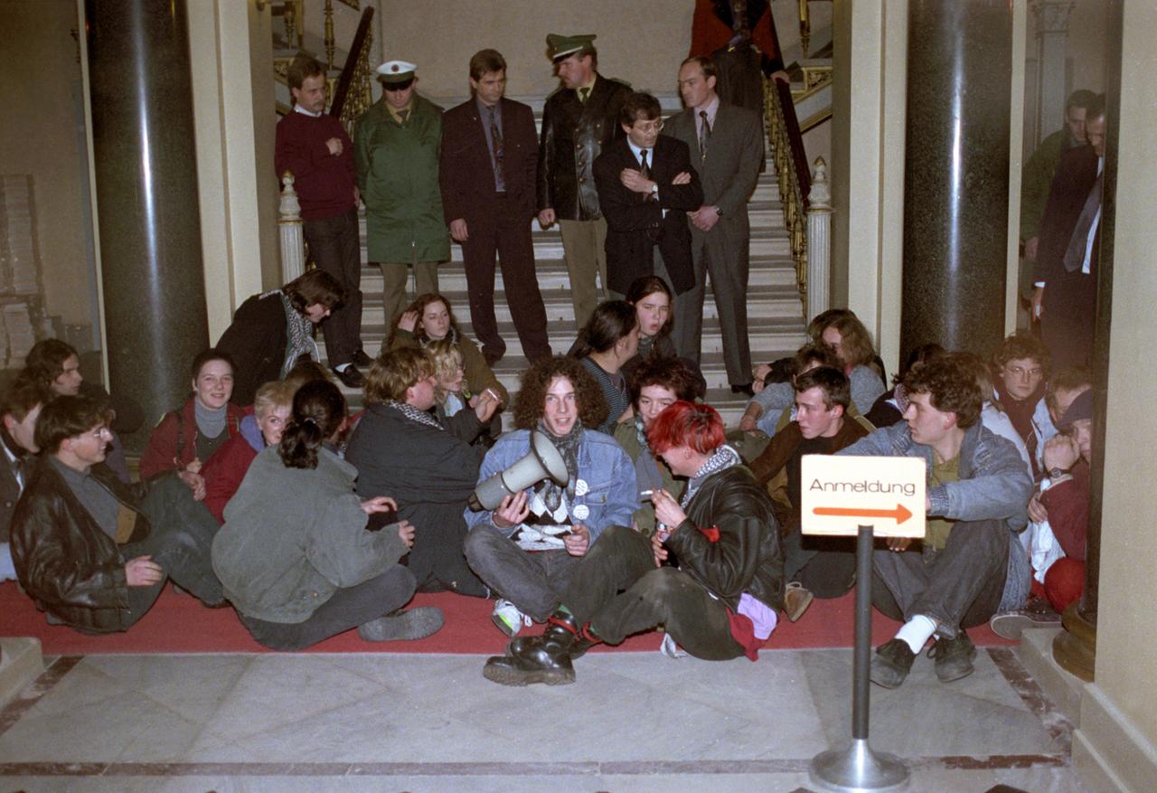 Knapp zwanzig Jugendliche sitzen in einem repräsentativen Gebäude auf dem Boden vor einer Treppe. Auf den ersten Stufen der Treppe nach oben hinter ihnen stehen zwei Polizisten in Uniform und mehrere Männer im Anzug mit Krawatte. Einer der Jugendlichen hält ein Megaphon in der Hand.