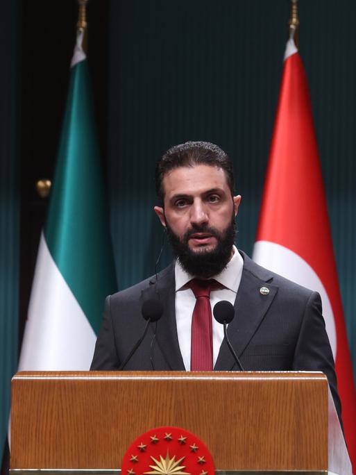 Ahmed al-Sharaa, der neue Machthaber in Syrien, steht während einer Pressekonferenz hinter einem Sprechpult. Dahinter die syrische und die türkische Flagge.