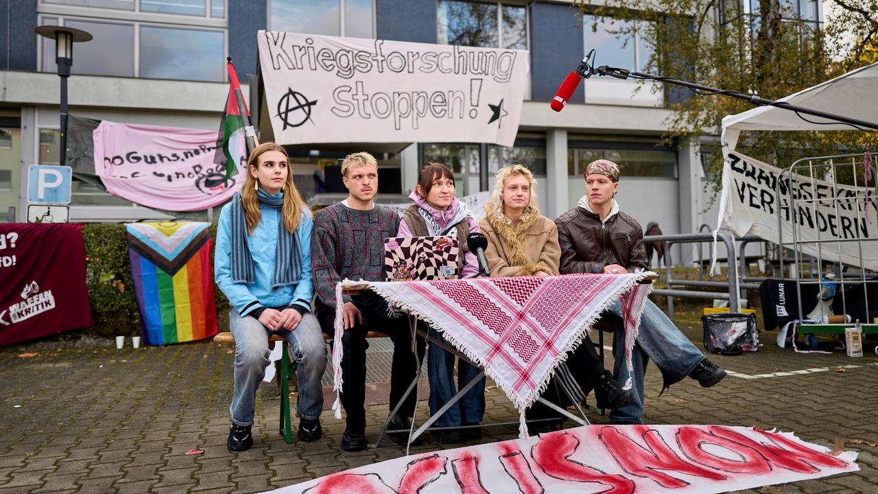 Fünf Studenten sitzen vor einem Unigebäude an einem Tisch, über den eine Kufiya ausgebreitet wurde. Ihnen wird ein Mikrofon entgegengehalten. Fünf Studenten sitzen vor einem Unigebäude an einem Tisch, über den eine Kufiya ausgebreitet wurde. Ihnen wird ein Mikrofon entgegengehalten.