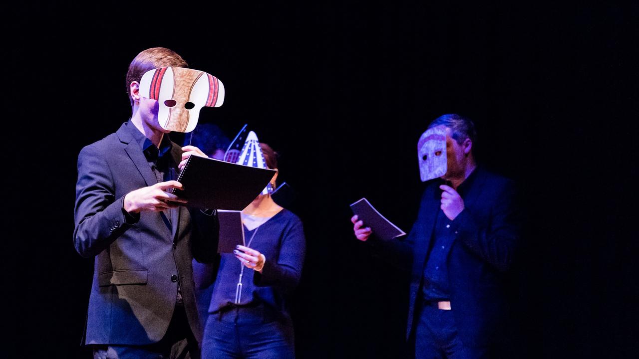 Drei Sänger und Sängerinnen gehen durch einen dunklen Raum und halten Masken vor ihre Gesichter.