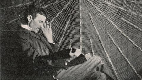 Nikola Tesla, im Profil, sitzt auf einem Stuhl und liest in einem Buch, er sitzt vor einer großen Spiralspule.