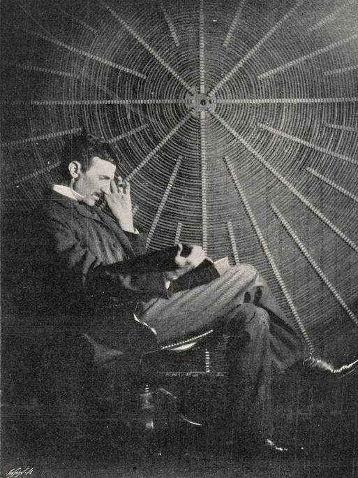Nikola Tesla, im Profil, sitzt auf einem Stuhl und liest in einem Buch, er sitzt vor einer großen Spiralspule.