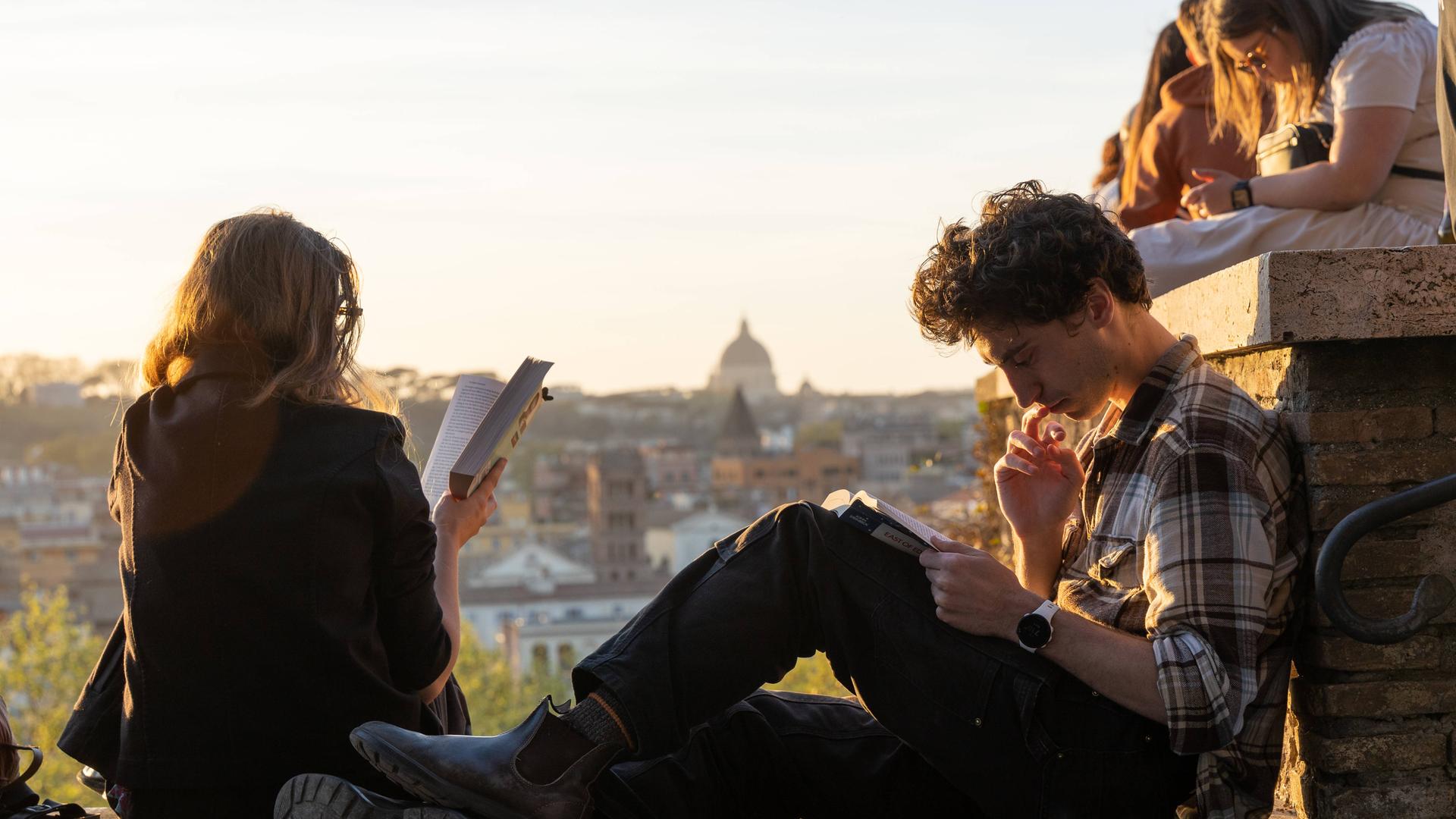 Eine junge Frau und ein junger Mann sitzen Bücher lesend auf einer Mauer im Orangengarten in Rom, mit dem Petersdom und Sonnenuntergang im Hintergrund