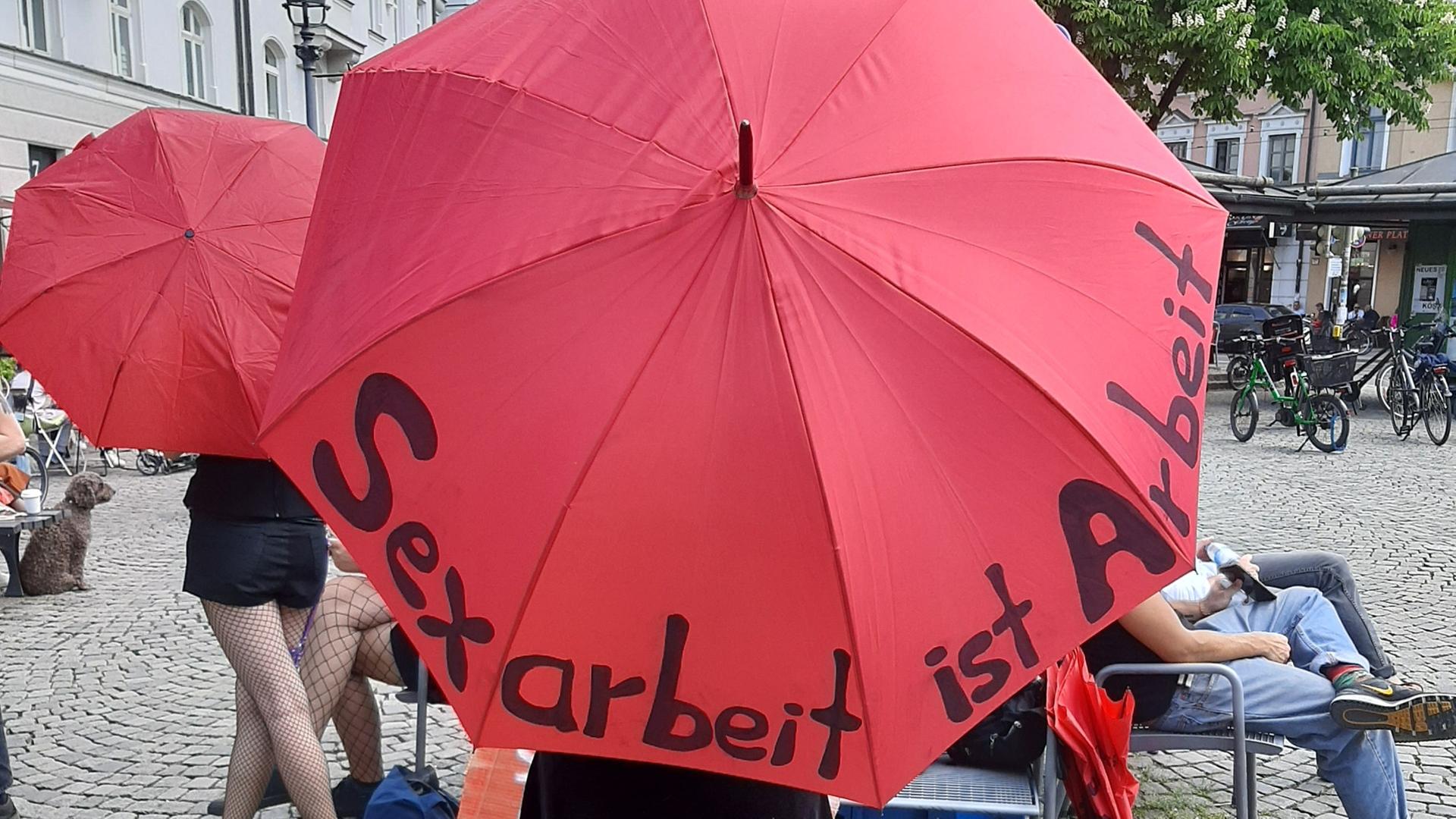 Eine Frau hat bei einem Pressetermin des Bundesverbandes Sexuelle Dienstleistungen e.V. einen roten Schirm über sich, auf dem "Sexarbeit ist Arbei" steht. 