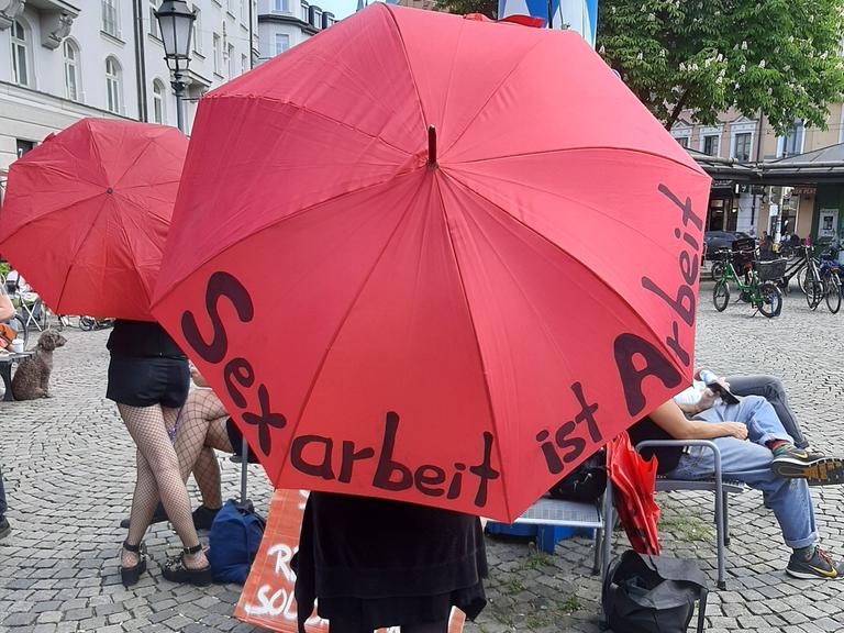 Eine Frau hat bei einem Pressetermin des Bundesverbandes Sexuelle Dienstleistungen e.V. einen roten Schirm über sich, auf dem "Sexarbeit ist Arbei" steht. 