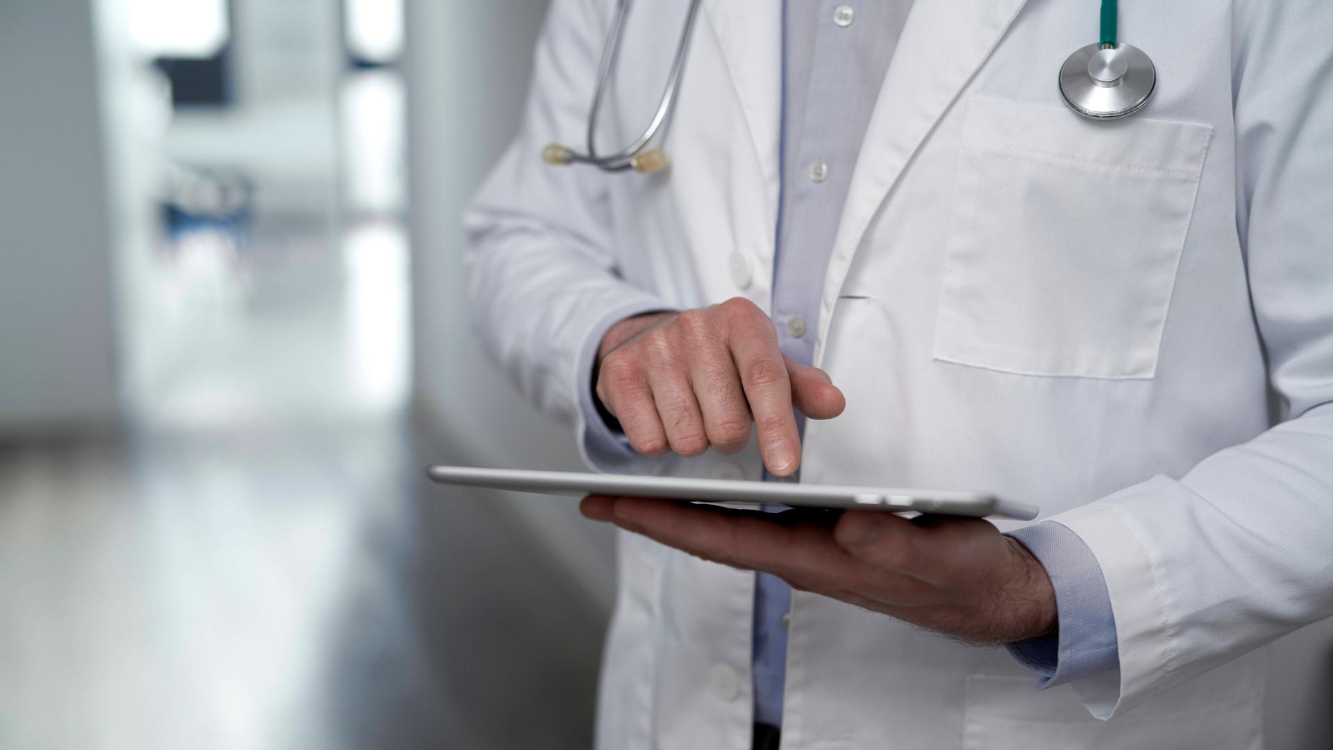 Symbolfoto: Mann in Arztkittel und Stetoskop berührt ein Tablet mit seinem Zeigefinger.