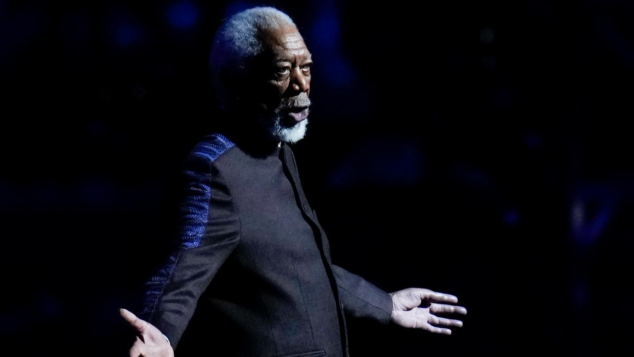 Freeman im dunklen Jackett steht auf einer Bühne, breitet die Arme aus und sagt etwas. Der Hintergrund ist schwarz. Er ist nur schwach beleuchtet. 