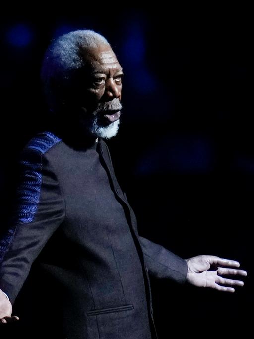 Freeman im dunklen Jackett steht auf einer Bühne, breitet die Arme aus und sagt etwas. Der Hintergrund ist schwarz. Er ist nur schwach beleuchtet. 