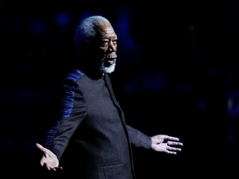 Freeman im dunklen Jackett steht auf einer Bühne, breitet die Arme aus und sagt etwas. Der Hintergrund ist schwarz. Er ist nur schwach beleuchtet. 