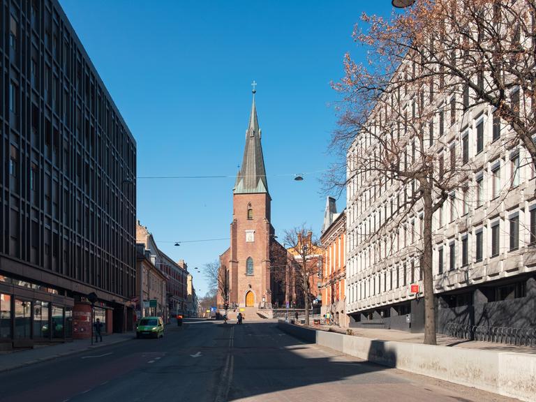 Straßenzug in Oslo, an dessen Ende eine von der Sonne beschienene Domkirche aus rotem Gestein steht.