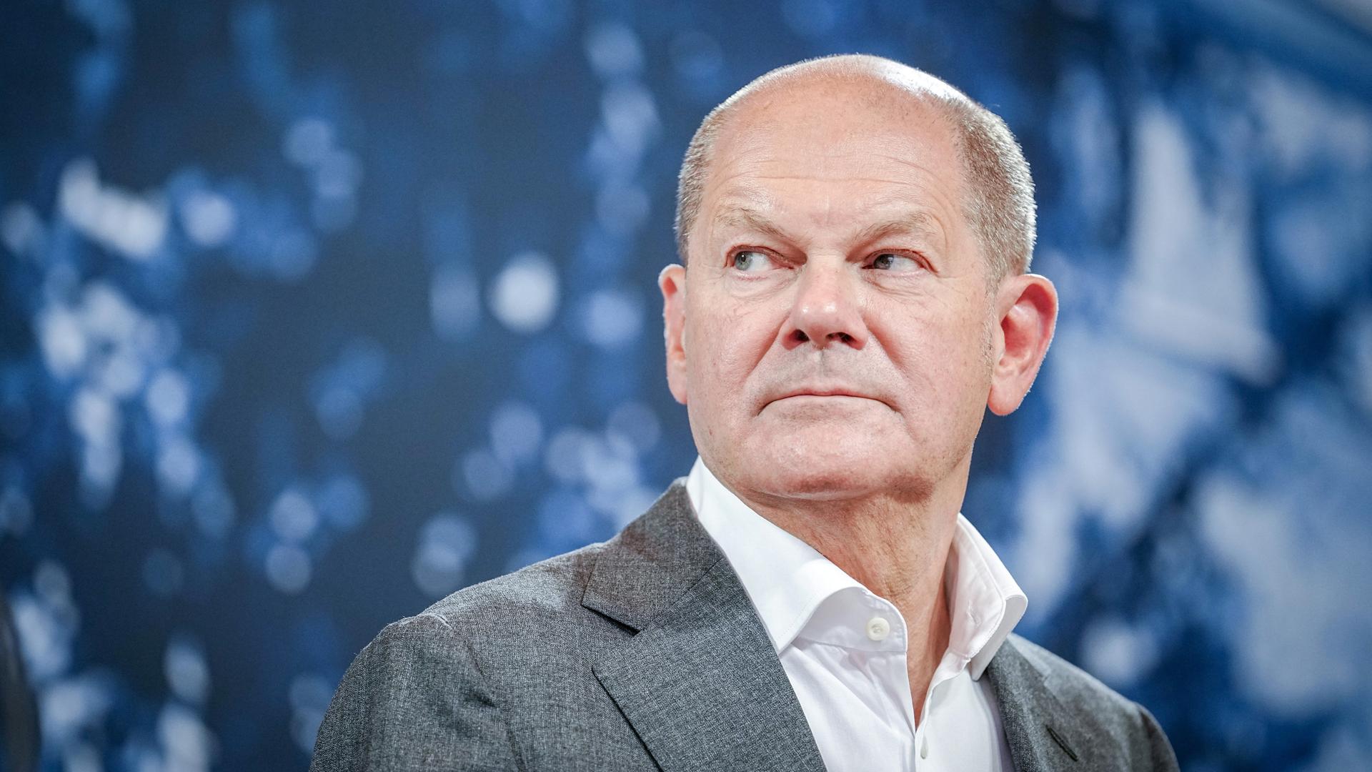 Bundeskanzler Olaf Scholz (SPD) sitzt vor blauem Hintergrund und schaut zur Seite.