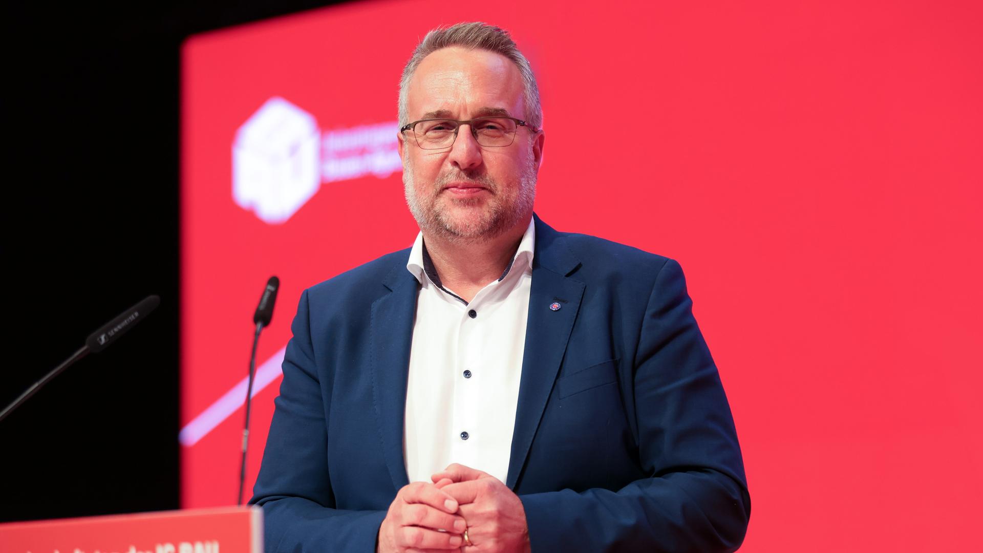 Carsten Burckardt im dunkelblauen Jackett steht vor einer roten Wand mit dem Gewerkschaftslogo und blickt leicht lächelnd in die Kamera. Er trägt eine Brille und hat einen Dreitage-Bart. Carsten Burckardt im dunkelblauen Jackett steht vor einer roten Wand mit dem Gewerkschaftslogo und blickt leicht lächelnd in die Kamera. Er trägt eine Brille und hat einen Dreitage-Bart.