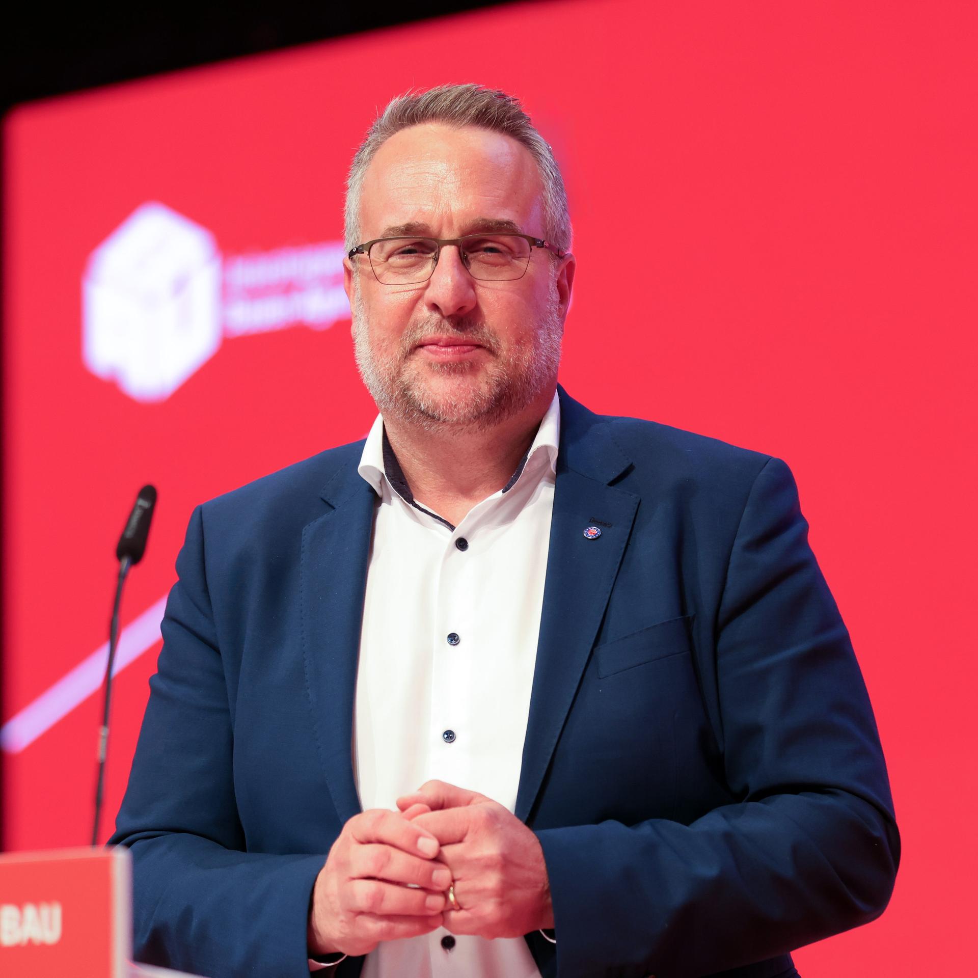 Carsten Burckardt im dunkelblauen Jackett steht vor einer roten Wand mit dem Gewerkschaftslogo und blickt leicht lächelnd in die Kamera. Er trägt eine Brille und hat einen Dreitage-Bart. 