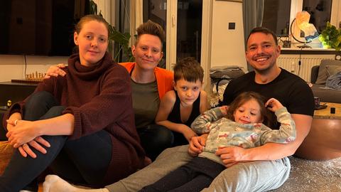 Eine Familie bestehend aus  zwei Müttern, einem Vater und zwei Kindern sitzen gemeinsam auf dem Wohnzimmerfußboden und lachen in die Kamera.