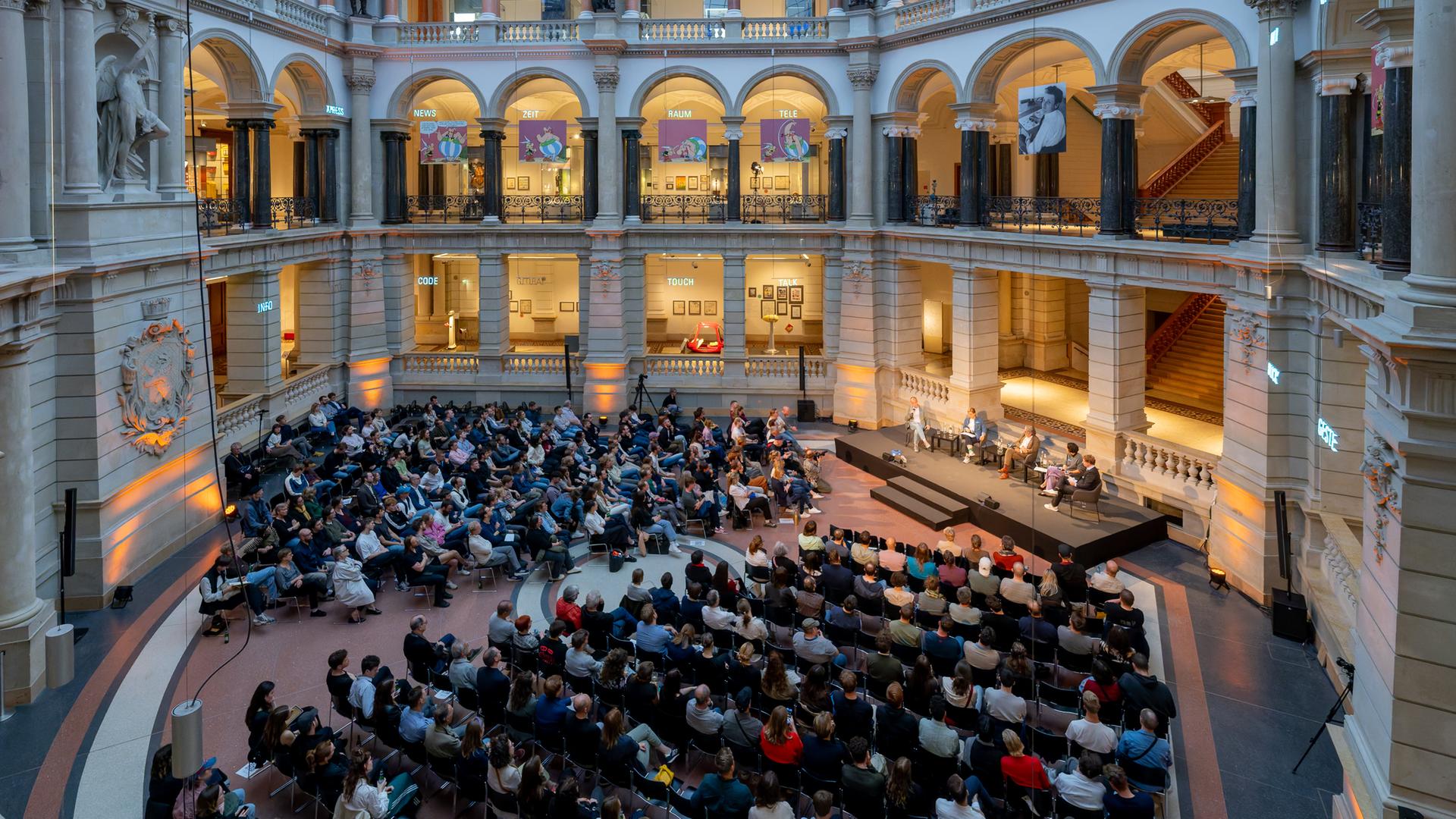 Blick von oben auf das 400köpfige Publikum und eine Bühne im Lichtsaal des Berliner Museums für Kommunikation, umrandet von Galerien und Säulen Blick von oben auf das 400köpfige Publikum und eine Bühne im Lichtsaal des Berliner Museums für Kommunikation, umrandet von Galerien und Säulen
