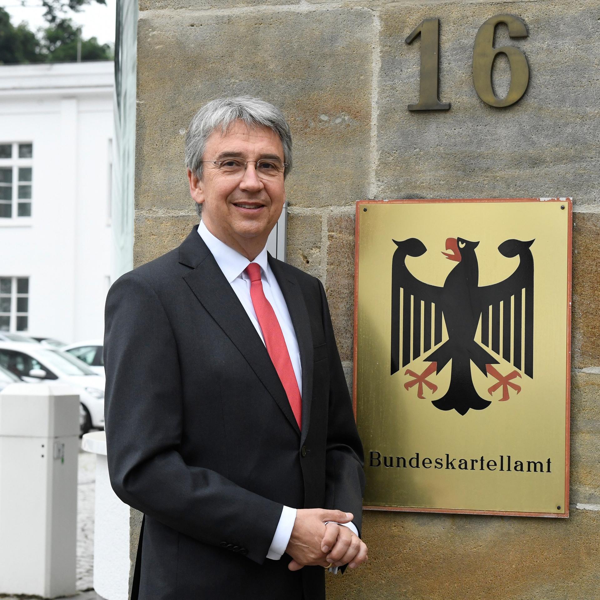 Andreas Mundt ist Präsident des Bundeskartellamtes. Er steht vor dem Sitz seiner Behörde und einem Schild mit Bundesadler. Er trägt einen schwarzen Anzuig, rote Kravatte zum weißen Hemd und hat graue Haare. 