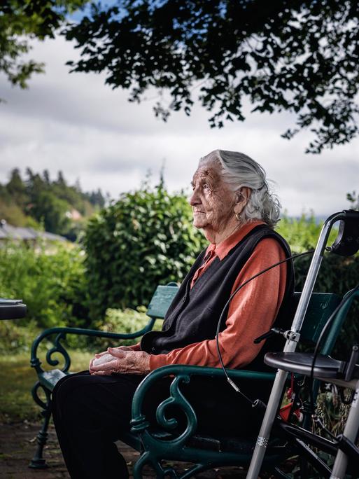 Alte Dame sitzt nachdenklich auf Parkbank, daneben ihr Rollator.